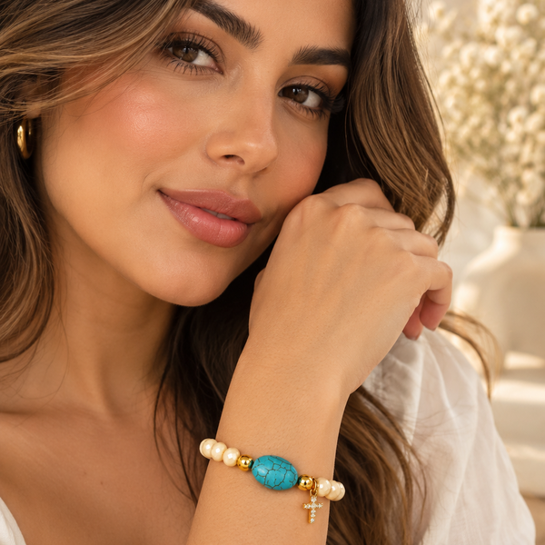 Descubre la pulsera Turquesa Celeste con cuentas color crema, piedra central turquesa y dije de cruz dorada. Un accesorio femenino y elegante